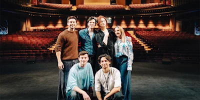 Six people pose together on a theater stage with empty seats and balconies visible in the background.