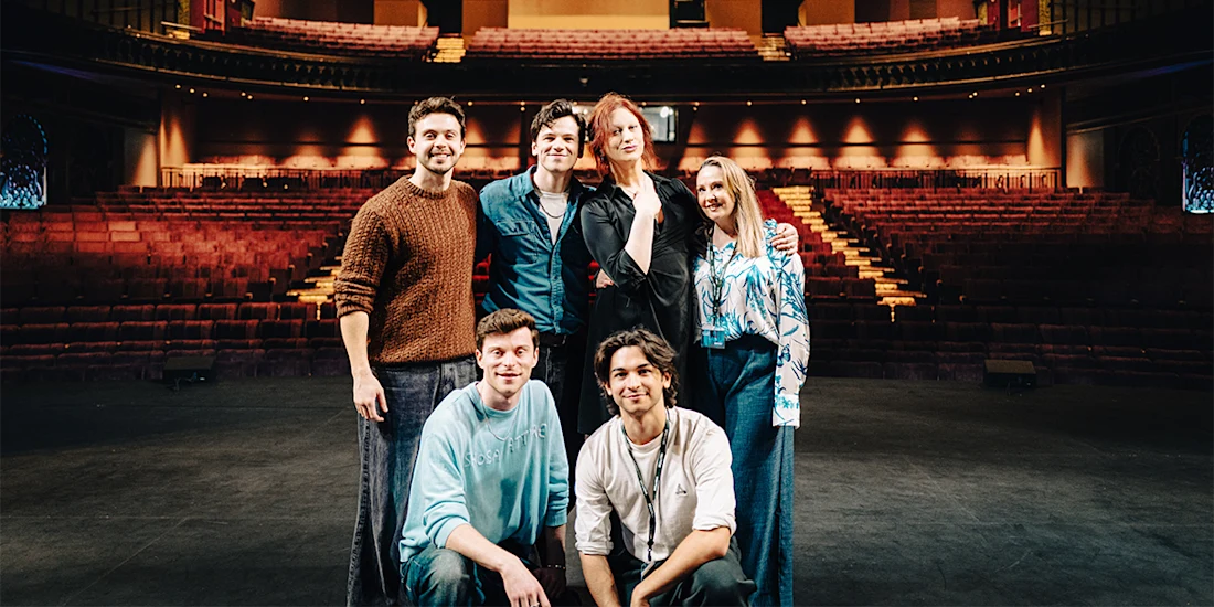 Six people pose together on a theater stage with empty seats and balconies visible in the background.