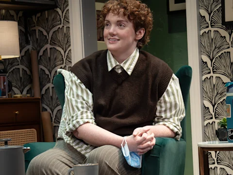 A person with short, curly reddish-brown hair, wearing a striped shirt and brown sweater vest, sits in a dark green armchair.