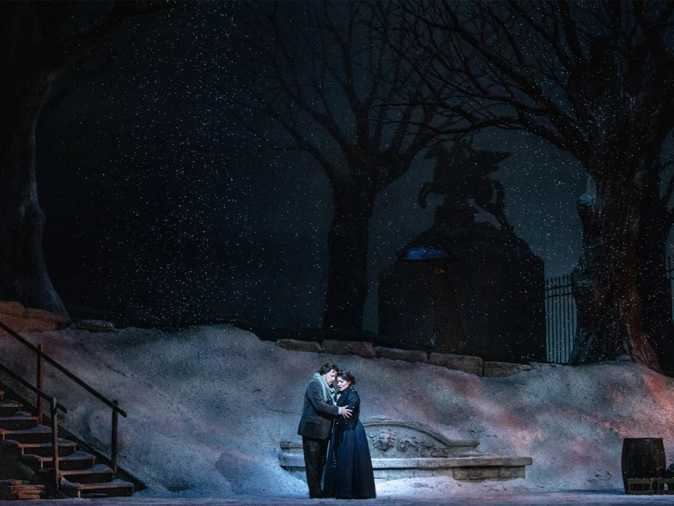 A couple stands close together in a snowy, dimly lit outdoor setting at night, near a stone structure and barren trees, with a statue in the background.