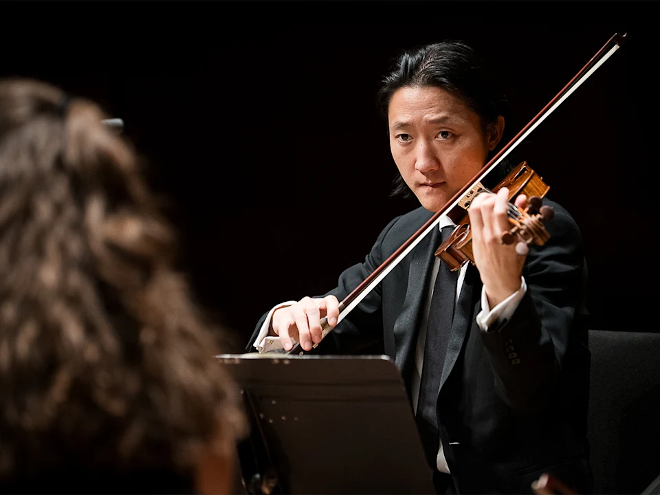 A violinist in a suit plays the violin with focus during a performance. Another person is partially visible in the foreground.