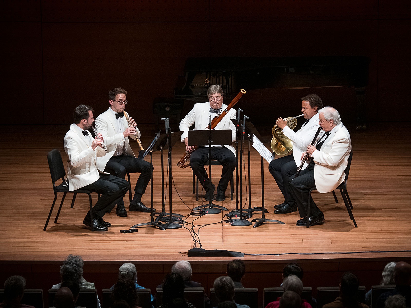 Five musicians in white jackets play wind instruments on stage in front of an audience, with music stands and microphones arranged in front of them.
