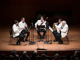 Five musicians in white jackets play wind instruments on stage in front of an audience, with music stands and microphones arranged in front of them.
