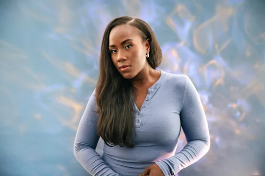 A woman with long dark hair wearing a long-sleeved light blue henley shirt against a blue and gold abstract background.