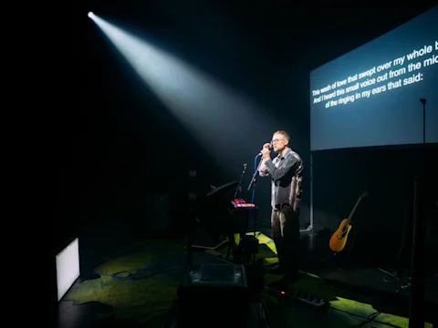A man stands on a stage performing with a microphone in front of a screen displaying song lyrics.