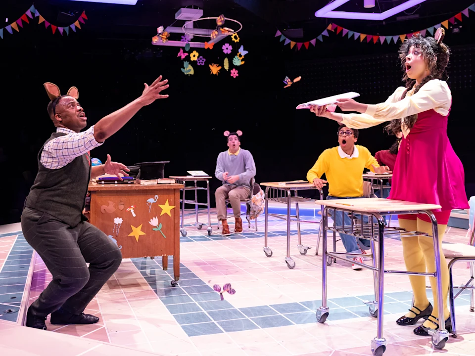 Four actors dressed as animals perform in a classroom set; one actor in a red dress throws papers to another who reaches out to catch them, while two others watch from desks.