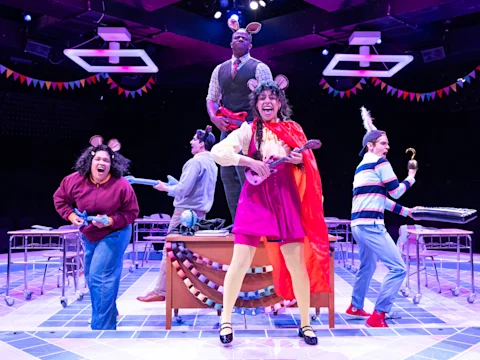 Five actors in colorful costumes with mouse ears perform energetically on stage, holding props and musical instruments, with festive decorations and classroom desks in the background.