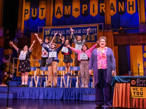 Stage scene from a spelling bee event with six participants holding number cards and a presenter in front. A banner reads "Putnam County Spelling Bee.