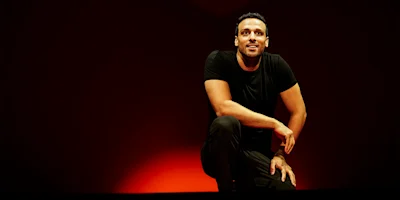 A man in a black t-shirt kneels on one knee on stage against a dark red background, looking upwards and smiling.