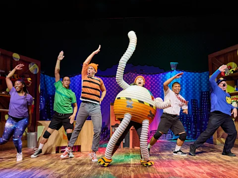 Six performers in colorful costumes dance energetically on a stage set resembling a child's playroom, with a character in a large striped circular outfit at the center.