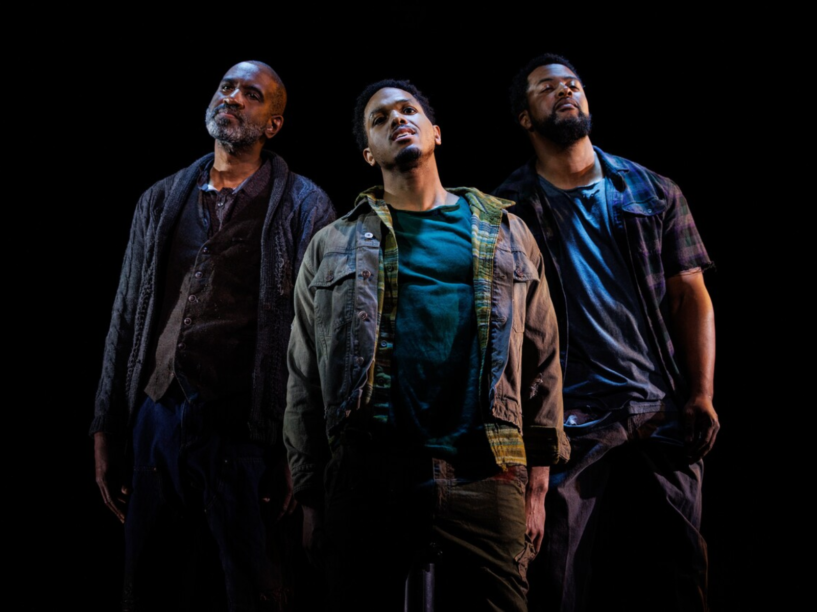 Three men stand close together in dim lighting against a black background, all looking slightly upwards with serious expressions.