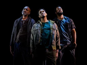 Three men stand close together in dim lighting against a black background, all looking slightly upwards with serious expressions.