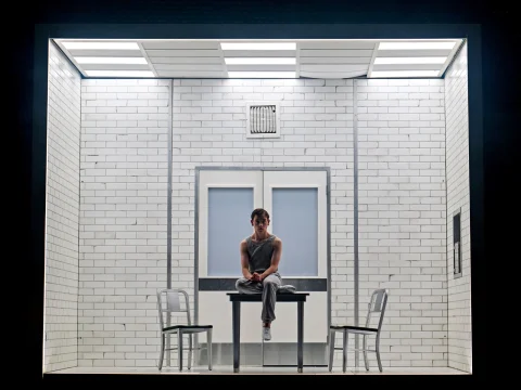 Louis McCartney as Henry Creel in Stranger Things: The First Shadow sits on a table in a minimalist white room with tiled walls, two chairs, and fluorescent lighting overhead.