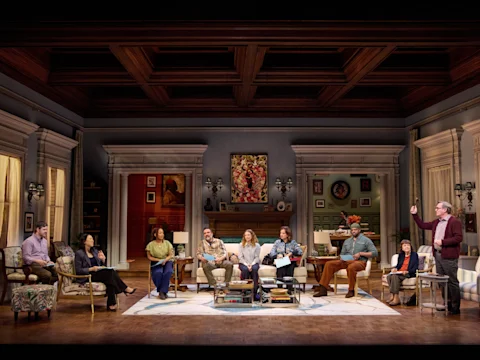 A group of people sit in a large, well-furnished living room set on stage, engaged in conversation, with one person standing and gesturing.
