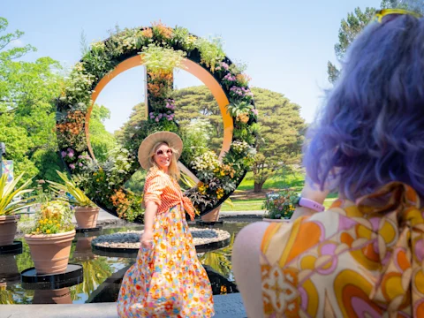 A woman in a colorful dress and sun hat poses smiling in front of a large floral peace sign, while another person with purple hair takes her photo.