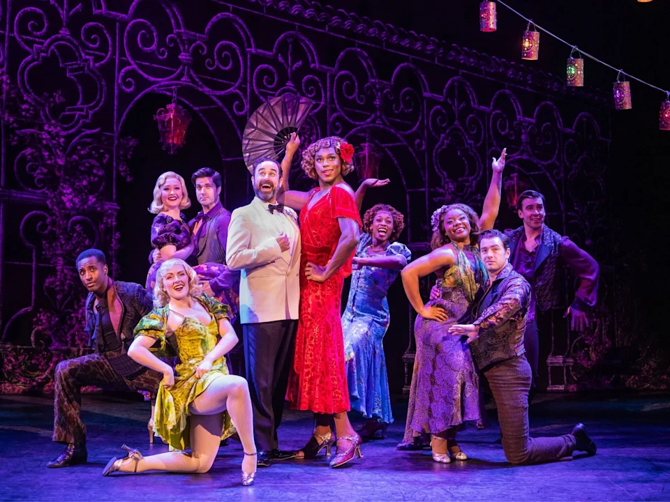 Performers in vibrant costumes pose energetically on stage, with decorative purple backdrop and colorful hanging lights.