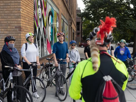 A group of cyclists wearing helmets gather on a sidewalk near a brick building with colorful art on the wall. One cyclist, seen from the back, appears to be addressing the group.