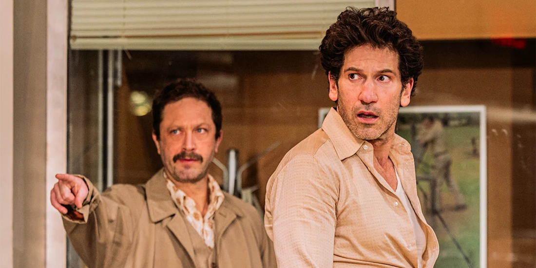 Two men in beige shirts stand indoors; one in the foreground looks surprised, while the other in the background points outward with a serious expression.