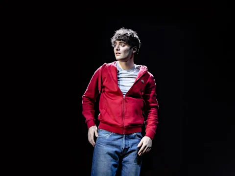 A young man in a red hoodie stands alone in a spotlight, looking troubled.