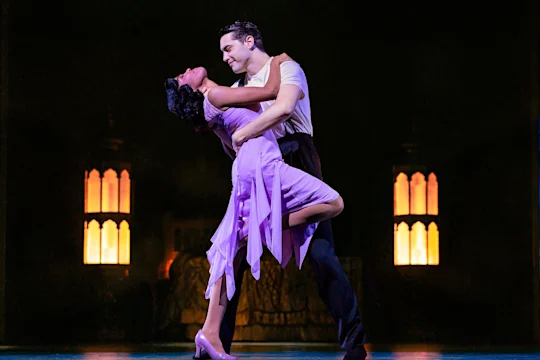 A man and woman in formal attire perform a dramatic dance pose on stage with warm yellow lights in the background.