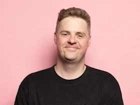 A person with short light hair wearing a black t-shirt stands in front of a plain pink background, smiling slightly at the camera.