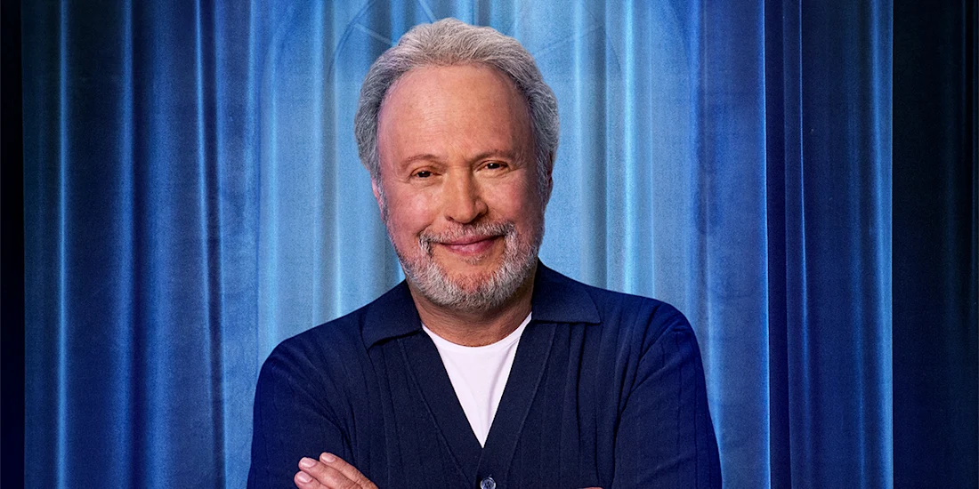 A man with gray hair and a beard stands in front of blue curtains, smiling and crossing his arms, wearing a dark blue shirt over a white T-shirt.