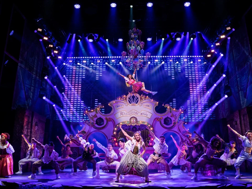 A vibrant stage performance featuring a group of actors in colorful costumes, with one actor suspended in the air. The backdrop includes a large sign reading "Renaissance Ball" and elaborate lighting effects.