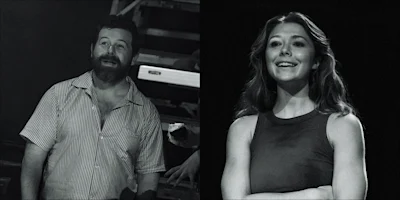 A man with a beard in a striped shirt and a woman in a sleeveless top stand under stage lighting, both looking slightly upward and smiling.