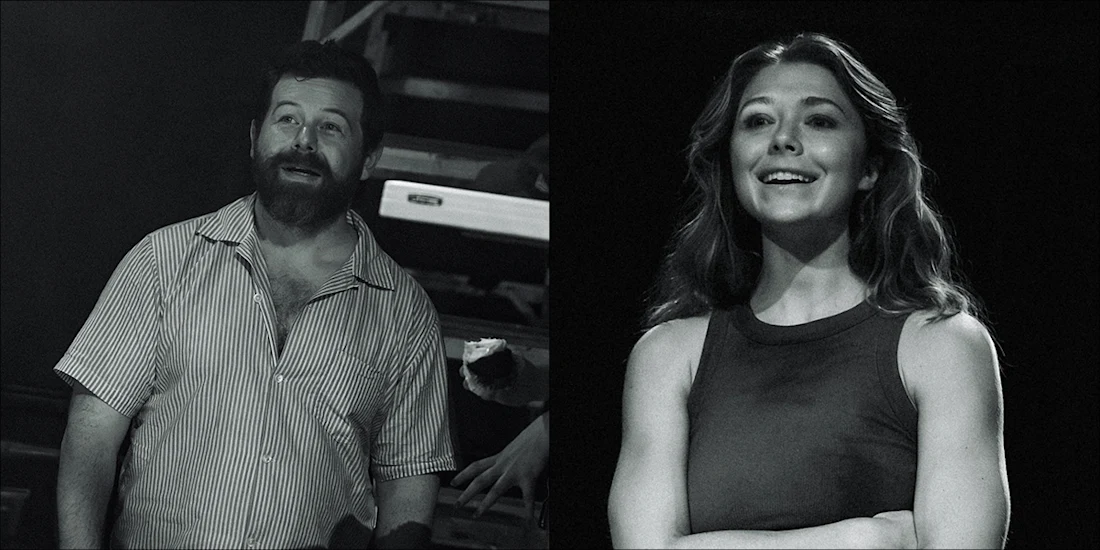 A man with a beard in a striped shirt and a woman in a sleeveless top stand under stage lighting, both looking slightly upward and smiling.