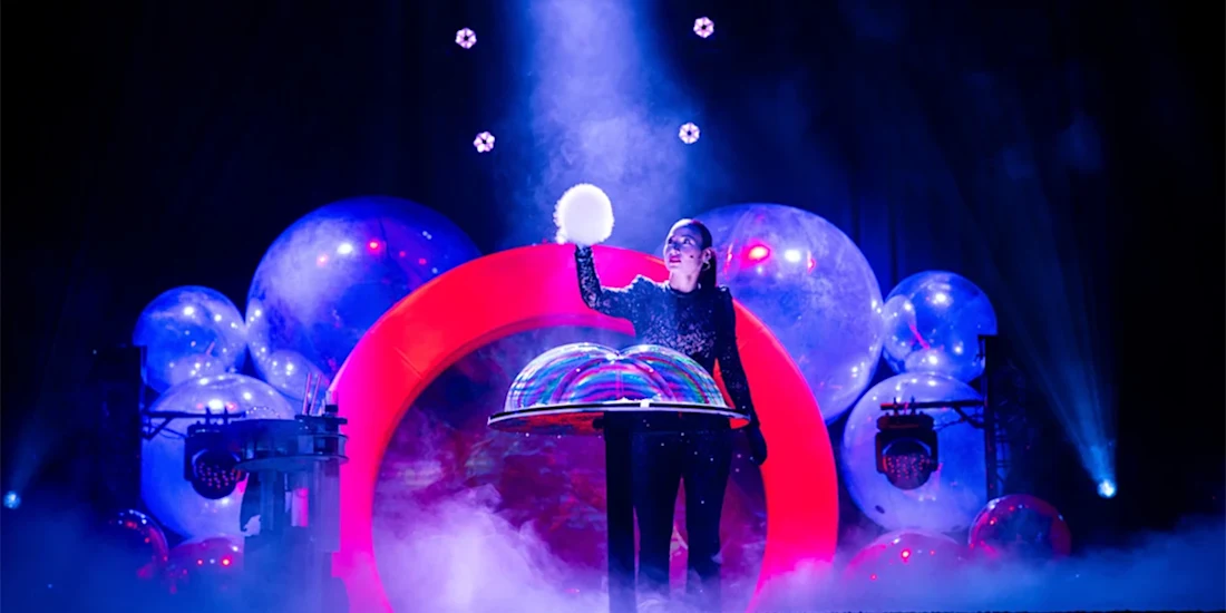 A performer in a sparkly outfit stands on stage surrounded by large bubbles and colorful lights, holding a glowing orb under a spotlight with fog at their feet.