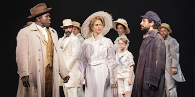 A group of people in period costumes stand together on stage, with a woman in a white dress and hat at the center, flanked by two men facing each other.