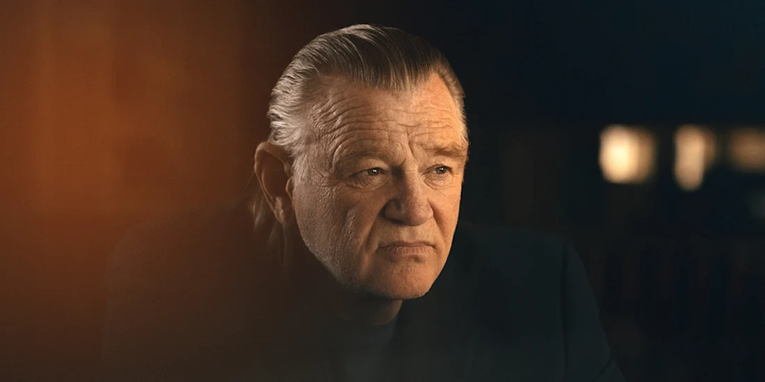 An older man with slicked-back gray hair and a serious expression sits indoors in low, warm lighting.