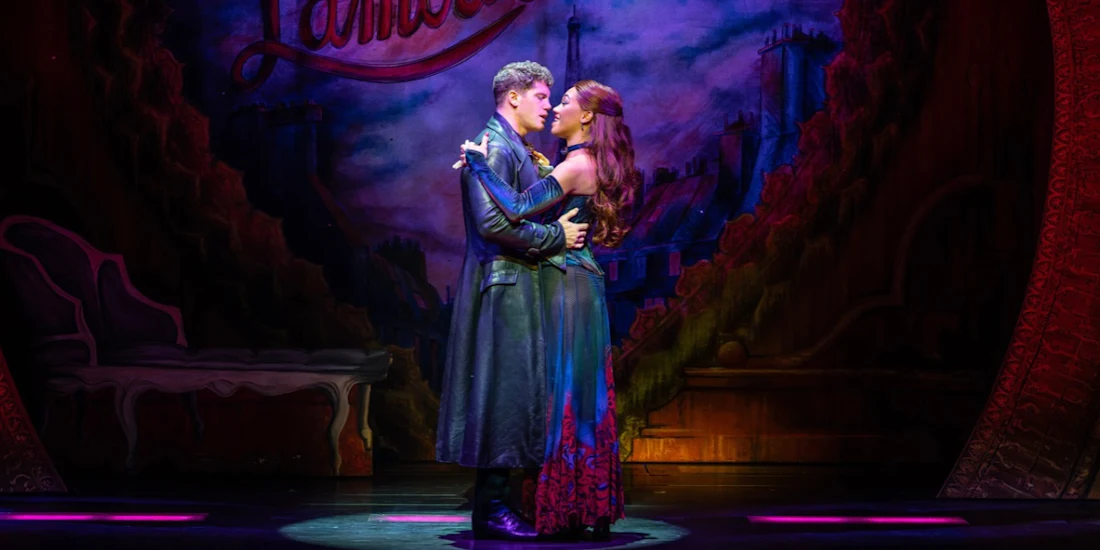 A man and woman in theatrical costumes embrace on stage in front of a painted backdrop of Paris.