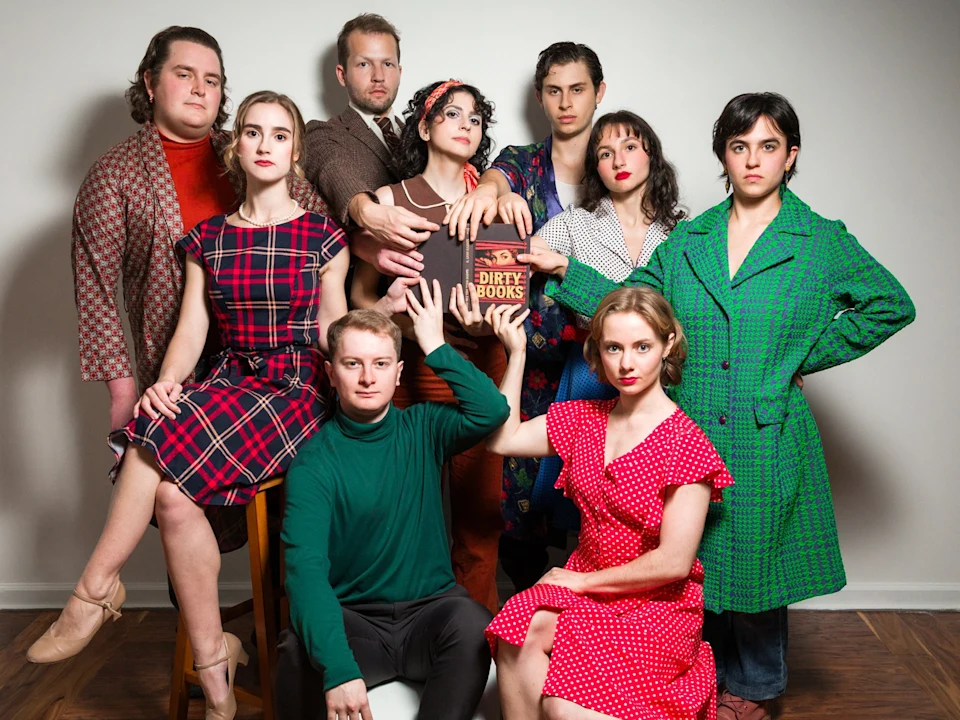 Eight people pose indoors against a blank wall, with two holding a brown book labeled "Dirty Books." They wear colorful, vintage-inspired clothing and look toward the camera.