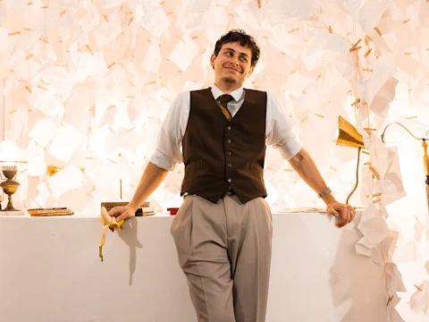 A man in a vest and tie leans against a white counter, holding a banana, with a background covered in scattered sheets of paper.