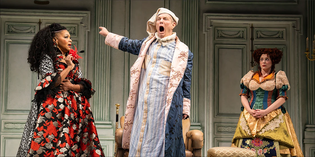 A woman in a red and black patterned dress gestures toward a man in a nightcap and robe who is shouting, while another woman in a historical gown stands to the right.