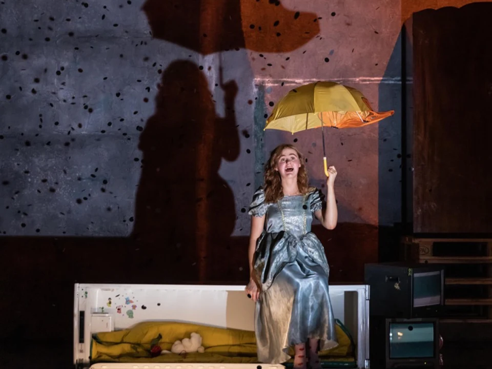 A woman in a long dress sits on a bed holding an open yellow umbrella, casting a shadow on the wall behind her.