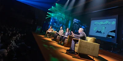 Four people sit on stage having a panel discussion in front of an audience, with a cartoon projected on a screen behind them and colorful stage lights overhead.