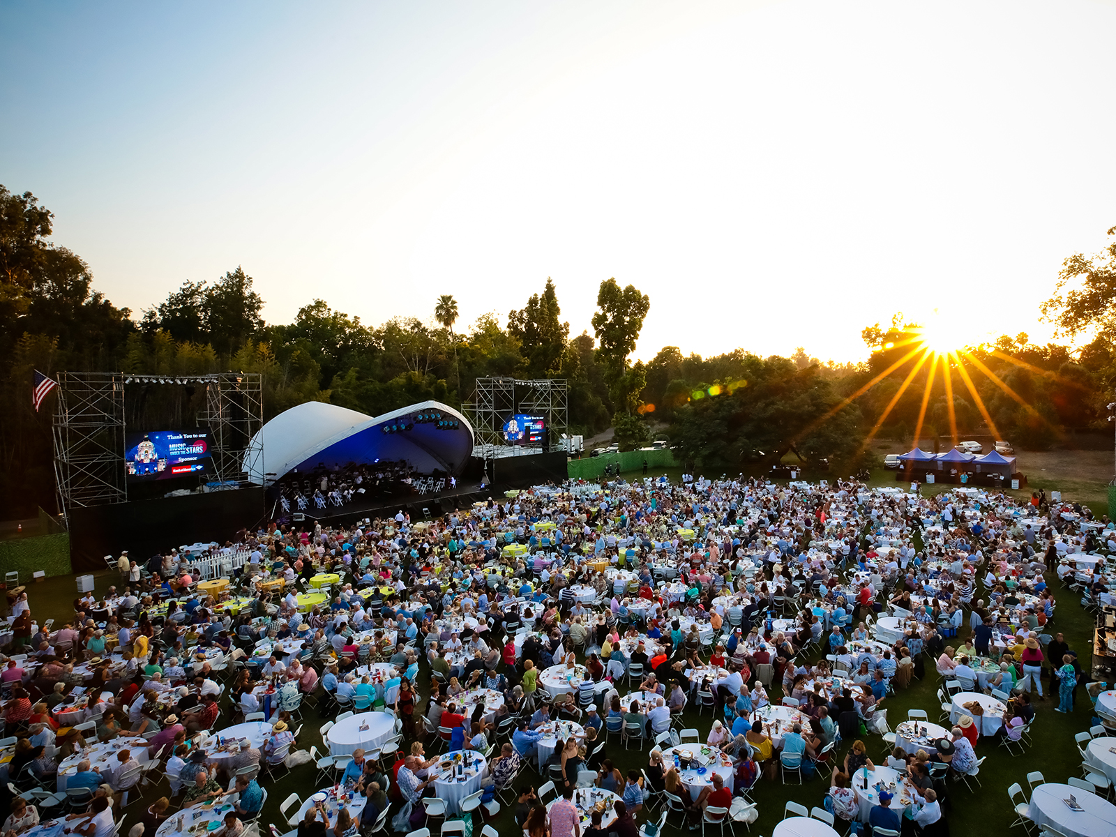 A large crowd attends an outdoor music concert as the sun sets. The audience sits at tables, and a stage with a band is visible. 