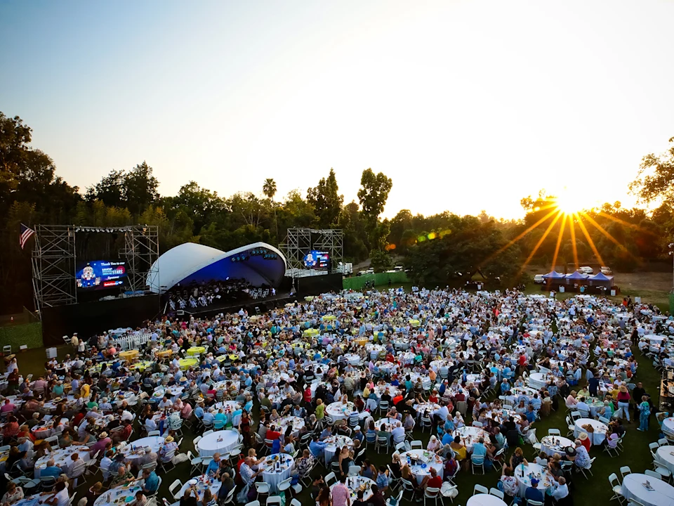 A large crowd attends an outdoor music concert as the sun sets. The audience sits at tables, and a stage with a band is visible. 