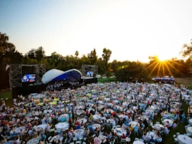 A large crowd attends an outdoor music concert as the sun sets. The audience sits at tables, and a stage with a band is visible.