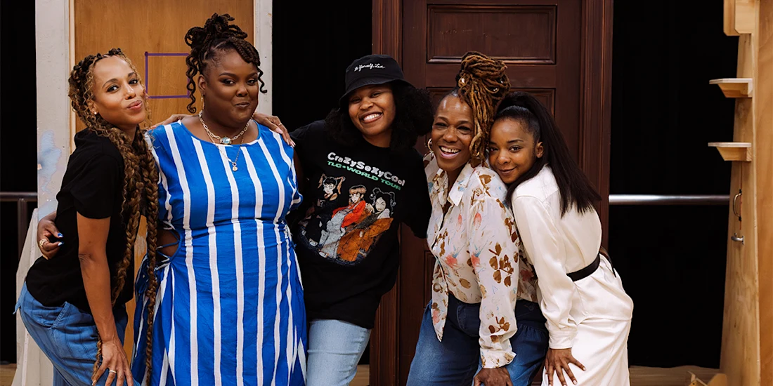 Five women stand closely together, smiling at the camera, in front of a backdrop featuring a wooden door and stairs.