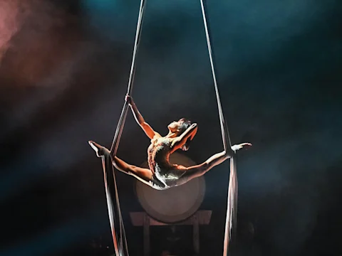 An aerial silk performer in a split pose, suspended midair, with dramatic stage lighting and a drum in the background.