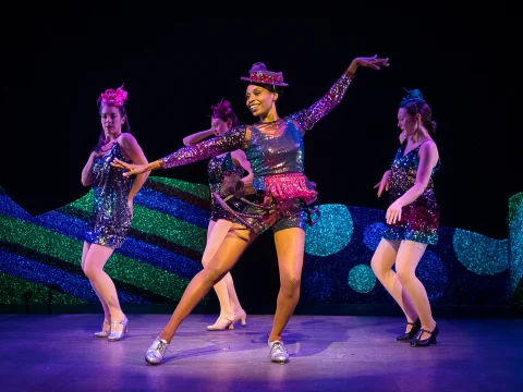 Four dancers perform on stage in colorful, sequined costumes and hats. One dancer in the center wears bright pink and purple and strikes an expressive pose. A stylized, colorful background is visible.