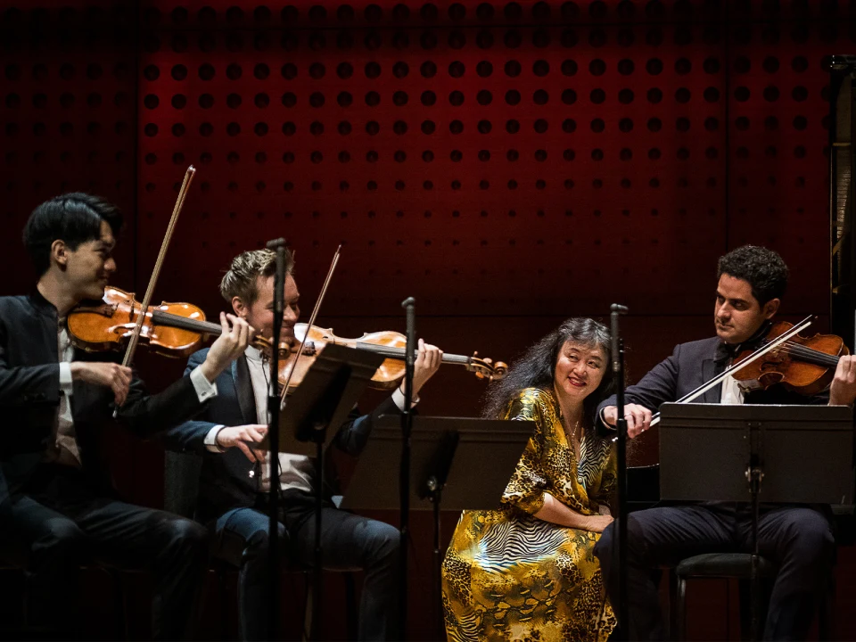 A string quartet performs on stage, with two violinists, a cellist, and a violist. The setting is a warmly-lit auditorium with a perforated red backdrop. The musicians are focused on their instruments.