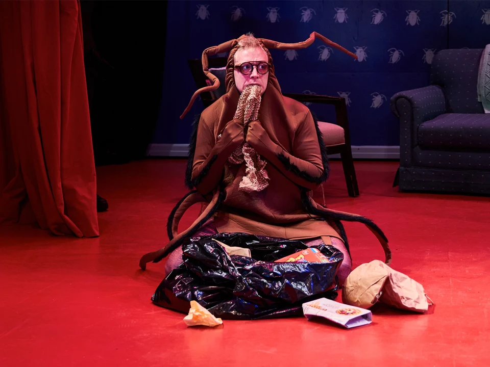 Person in insect costume sits on red floor, eating a wrapped item, surrounded by crumpled bags and discarded packages.