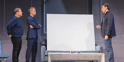 Three men stand around a white blank canvas in a modern room; two are on one side with arms crossed, and one is on the opposite side holding an object.