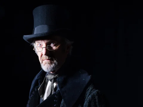 Older man in a top hat and glasses, wearing a dark coat, against a black background.