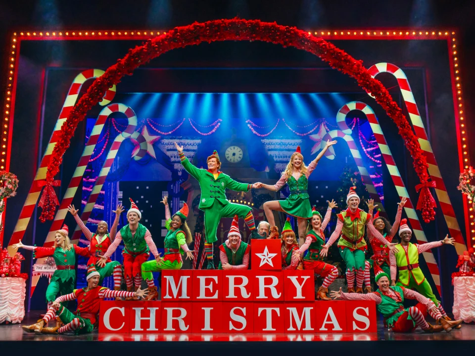 Performers dressed as elves and other Christmas-themed characters pose in front of a large stage in Elf the Musical on Broadway.