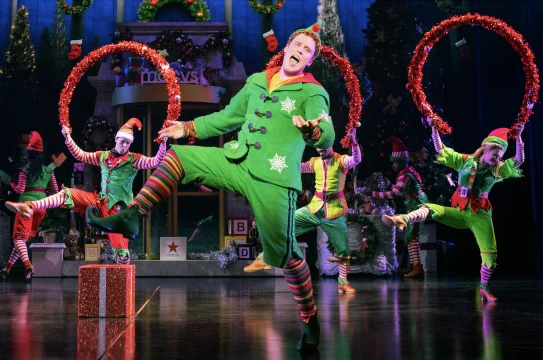 Performers dressed as elves and other Christmas-themed characters pose in front of a large "Merry Christmas" sign on stage, accompanied by festive decorations including candy canes and red garlands.